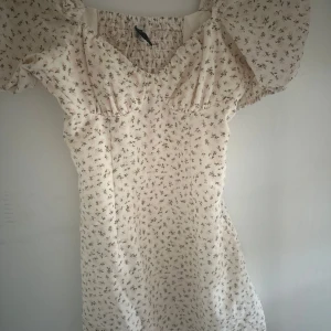 Blommig kortklänning från H&M Divided - Supersöt ljus kortklänning från H&M Divided med små blommor i beige och rosa. Klänningen har puffiga kortärmar, smockad rygg och v-ringad hals. Perfekt för sommarens alla tillfällen, med mjukt och lätt bomullsmaterial som känns skönt mot huden Tar frakt!.
