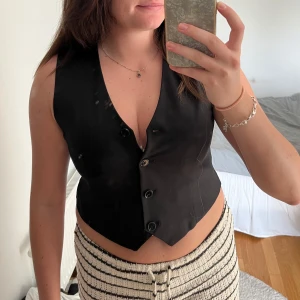 Svart klassisk väst med knappar - Säljer en svart väst med v-ringad hals och fyra knappar framtill. Västen har en klassisk, lite croppad passform som sitter snyggt över midjan. Perfekt att styla över en t-shirt eller skjorta för en cool look. Materialet känns slätt och lätt, troligen polyester.