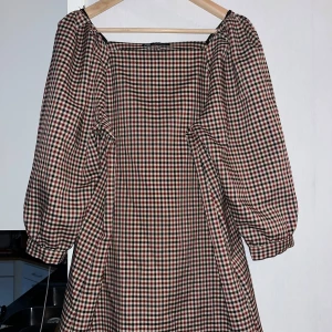 Rutig kortklänning med puffärm från Zara - Superfin rutig kortklänning från Zara med fyrkantig ringning och puffiga långärmar. Klänningen har en loose passform och är perfekt för dig som gillar vintagevibbar. Färgerna är en mix av rött, svart och vitt. Perfekt att styla med boots eller sneakers.