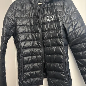 Svart pufferjacka från EA7 Armani - Snygg svart pufferjacka från EA7 Emporio Armani med quiltad design och dragkedja framtill. Jackan har en stilren look med logga på bröstet och är perfekt för kyligare dagar. Lätt och smidig att bära, passar till många outfits.