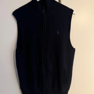 Mörkblå stickad väst från Ralph Lauren - Snygg mörkblå stickad väst från Ralph Lauren med dragkedja framtill och diskret broderad logga på bröstet. Västen har hög krage och två fickor på sidorna. Perfekt att slänga över en hoodie eller t-shirt. Ny pris för denna var 3200kr