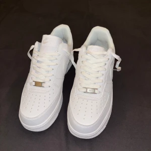 Nike Air Force 1 helvita sneakers - Helt nya Air Force 1, aldrig använda, klassiska Nike sneakers i helvitt skinn. Skorna har låg profil, rund tå, snörning och perforerade detaljer på tån för extra ventilation. Perfekta för dig som vill ha en clean och tidlös look till vardags.