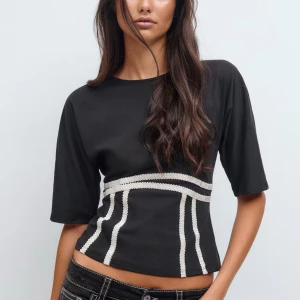 Svart topp med vita detaljer - Svart topp med halvkort ärm och vita, pärlliknande dekorativa linjer som ger en cool kontrast. Toppen har en croppad passform och rund hals. Perfekt för dig som gillar stilrena plagg med en twist.
