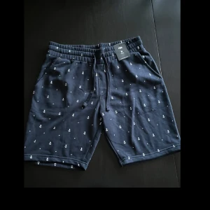 Marinblå shorts med ankare från FSBN - Snygga marinblå shorts från FSBN i storlek M, med vita ankare och prickar som mönster. De har elastisk midja med snörning, sidofickor och en bakficka. Mjukt bomullsmaterial som är perfekt för chill dagar och sommarhäng.