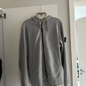 Grå hoodie/kofta från Stone Island - Säljer en grå hoodie från Stone Island med dragkedja, huva och kängurufickor. Klassisk logga på ärmen och skön passform. Bra skick. Passar både för storlek S/M. 