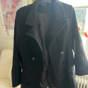 Svart rock från Capture - Stilren svart rock från Capture med dubbelknäppning och klassisk krage. Jackan har en rak passform och är helfodrad, perfekt för kyligare dagar. Snygg att slänga över en hoodie eller stickad tröja för en clean look.