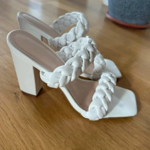 Vita sandaler med flätade remmar - Snygga vita sandaler med chunky klack och två breda, flätade remmar över foten. Skorna har fyrkantig tå och är tillverkade i ett skinnliknande material. Perfekta för dig som vill sticka ut med en trendig och stilren look.