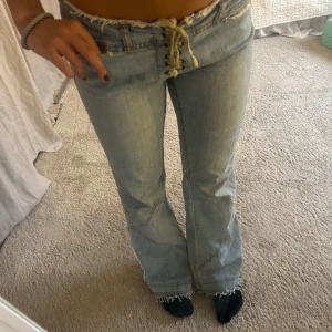Ljusblå bootcut jeans med snörning - Säljer ett par ljusblå jeans med bootcut passform och låg midja. Jeansen har snörning framtill istället för vanlig gylf, råa fransade kanter i midjan och nertill, samt coola sömdetaljer på benen. Lite stora på mig som är S/M så passar nog bäst på M
