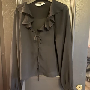 Svart blus med volang och knyt från Kappahl  - Supersnygg svart blus från Kappahl med volangdetaljer längs kragen och knyt framtill. Blusen är i ett tunt, lätt material och har långa ärmar med knyt vid ärmslutet. Perfekt för dig som gillar en romantisk och trendig look.💕💕