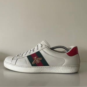 Gucci Ace sneakers med bi-detalj - Tja! Säljer ett par Gucci Ace sneakers i vitt skinn med klassiska gröna och röda ränder på sidan, och broderad guldfärgad bi. Sparsamt använda 7/10 skick.Skorna har snörning och kommer med original dustbag.