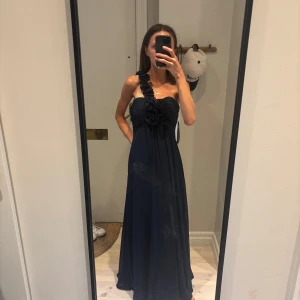 Marinblå balklänning med rosdetaljer - Säljer en marinblå balklänning med oneshoulder och rosdetaljer längs axelbandet. Klänningen har plisserad överdel och är tillverkad i ett lätt, flytande material med fodrad insida. Perfekt för bal eller fest, ger en elegant och modern look. OBS! Klänningen är enligt lappen i strl 4xs, men, passar en vanlig XS! Klänningen är köpt för ca 3000 kr.