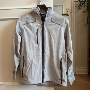 Ljusgrå vindjacka från Helly Hansen - Säljer en ljusgrå Helly Hansen Workwear-jacka. Jackan har huva, flera dragkedjeförsedda fickor och justerbara ärmslut. Perfekt för blåsiga dagar och snygg till streetwear. Materialet känns slitstarkt och jackan har en clean look med svarta detaljer. Är osäker på om modellen är till för kvinnor eller män , så valde unisex