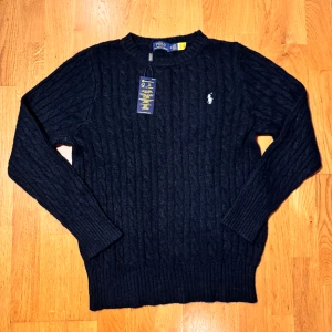 Mörkblå stickad tröja Polo Ralph Lauren - Kabelstickad mörkblå tröja från Polo Ralph Lauren med klassisk rund halsringning och broderad logga på bröstet. Tröjan har långa ärmar och ribbade muddar, perfekt för lager-på-lager under kyliga dagar.