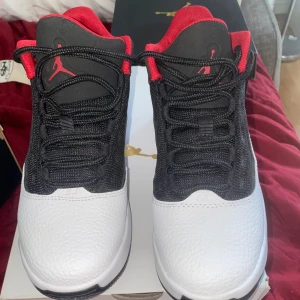 Svarta och vita Jordan sneakers - Snygga Jordan sneakers i svart och vitt med röda detaljer. Ovandelen är i svart mesh och syntet, medan tån och hälen är i vitt skinn. Röd insida och Jumpman-logga på plösen och sidan. Klassisk snörning och rund tå. Perfekta för dig som gillar streetstyle.