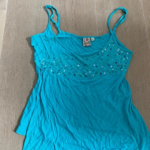 Turkos linnetopp med paljetter och pärlor - Säljer en turkos linnetopp med smala axelband. Toppen har meshparti upptill dekorerat med paljetter, pärlor och små stjärnor i olika färger. Perfekt för sommaren!