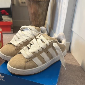 Beige Adidas Campus 00 - Snygga beige sneakers från Adidas med vita ränder på sidorna och vit sula. Skorna har breda vita skosnören och är i mocka med detaljer i syntet. Skorna har bara använts ett få tal gånger och är i super bra skick. 