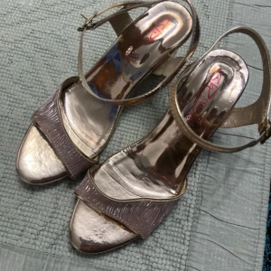 Guldiga sandaler med klack och rem - Säljer ett par snygga sandaler i metallic guld med bred rem över tårna och justerbar ankelrem. Skorna har öppen tå och klack, perfekt för att lyfta din outfit. Materialet är syntet med glansig finish.