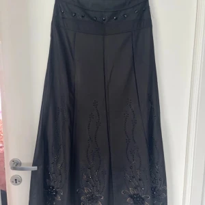 Svart långkjol med broderi och pärlor från Bolandi - Säljer en svart långkjol från Bolandi i storlek 38. Kjolen har vackra broderade detaljer och svarta pärlor längs nederdelen, vilket ger en elegant look. Materialet är glansigt och känns lyxigt. Perfekt för dig som vill sticka ut med en unik kjol. Är vintage från Iran.