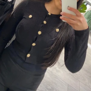 Svart kofta med guldknappar - Snygg svart kofta med dekorativa guldknappar framtill. Perfekt för att ge en elegant touch till din outfit. Koftan har långa ärmar och en figurnära passform.