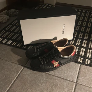 Gucci ace skor - Snygga svarta sneakers från Gucci med röd och blå rand samt en broderad bee-detalj på sidan. Strl 41,5, mycket bra skick, Originalboxen medföljer. Nypris 8000 kr, mitt pris 2399 kr. Vid funderingar eller frågor så är det bara att höra av sig!😀