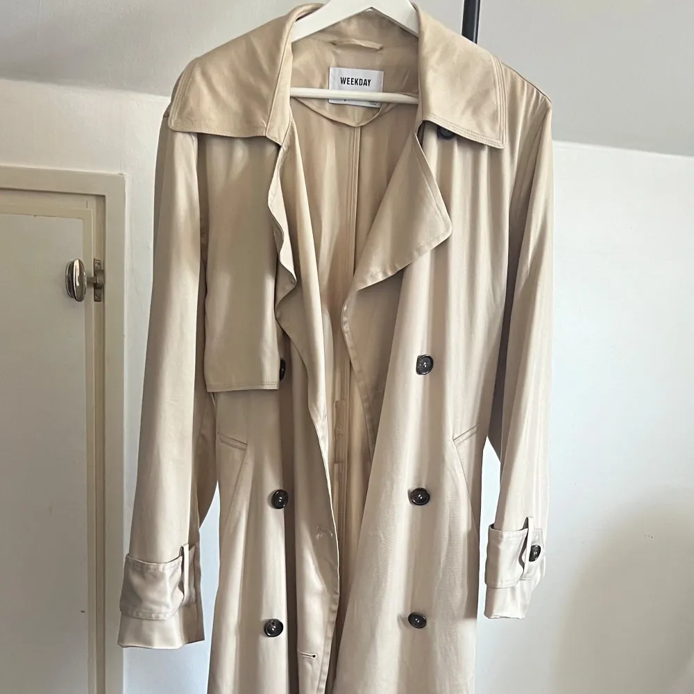 Snygg beige trenchcoat från Weekday med klassisk design. Jackan har knappar framtill och ett bälte i midjan för en justerbar passform. Perfekt för vår och höst!. Takit.