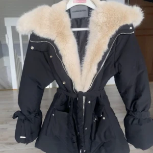 Svart parkajacka med fuskpäls från Marniére De Voir - Säljer en svart jacka med stor beige fuskpäls runt huvan. Jackan har dragkedja, knappar och bälte i midjan samt knytband vid ärmsluten. Perfekt för kyliga dagar och riktigt snygg detalj med pälsen.