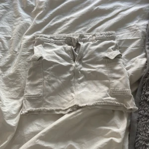 Vit jeanskjol från H&M - Säljer en vit jeanskjol från H&M med rå kant nertill och två stora fickor fram. Kjolen har dragkedja och knapp framtill samt bälteshällor. Perfekt för en avslappnad och trendig look.
