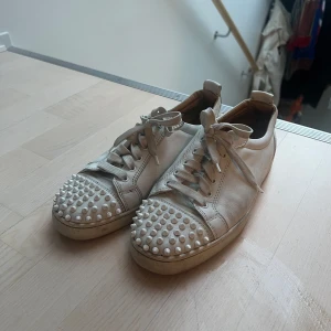 Christian Louboutin | Vita sneakers med nitar - Säljer ett par vita sneakers med coola nitar på tån och klassisk snörning. Skorna har en unik röd sula och är tillverkade i skinn. I använt skick. Storlek 42.