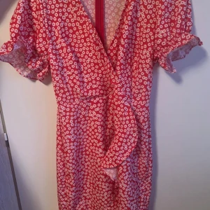 Röd blommig omlottklänning - Säljer en röd omlottklänning med vita blommor och korta puffärmar. Klänningen har v-ringning och volangdetaljer framtill. Perfekt för sommarens alla tillfällen!