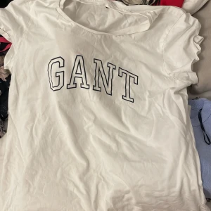 Vit t-shirt från GANT - Vit t-shirt från GANT med stor svart logotyp på bröstet. Klassisk rund halsringning och korta ärmar. Perfekt basic-plagg med enkel och stilren design.