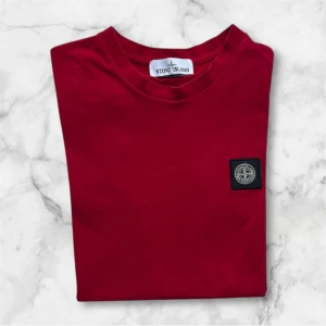 Röd t-shirt från Stone Island - Snygg röd t-shirt från Stone Island med klassisk rund hals och ikonisk logopatch på bröstet. Tillverkad i mjuk bomull som känns skön mot huden. Perfekt för dig som gillar stilrena och trendiga plagg.