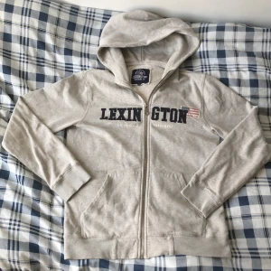 Lexington zip tröja - Säljer en ljusgrå hoodie från Lexington med dragkedja och huva. Tröjan har tryckt Lexington-logga och amerikansk flagga på bröstet samt två fickor framtill. Perfekt för en avslappnad stil. Orginal pris 1300