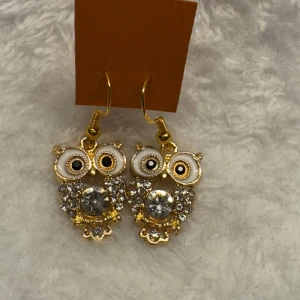 Guldiga uggleörhängen med strass - Säljer ett par unika örhängen i form av ugglor i guldton med vita detaljer och glittrande strass på kroppen. Perfekta för dig som vill sticka ut med ett lekfullt smycke!