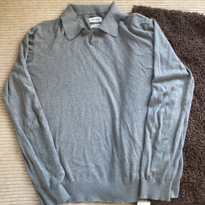 Grå silkeblandad pikétröja från These Glory Days - Säljer en stilren grå långärmad pikétröja från These Glory Days i silkeblandat material. Tröjan har klassisk krage och ribbade muddar vid ärmslut och nederkant. Perfekt för en clean och avslappnad look.