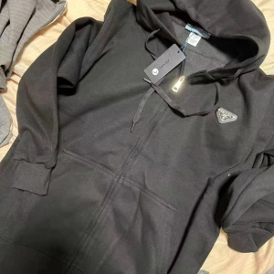 Prada jacka - Säljer en svart hoodie från Prada med dragkedja framtill och huva. Tröjan har en diskret Prada-logga på bröstet och ribbade muddar vid ärmslut och nederkant. Perfekt för en stilren och avslappnad look.