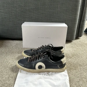 Svarta sneakers från Oliver Cabell med vit detalj - Säljer ett par svarta sneakers från Oliver Cabell med vit sula och vit häl samt en stor vit cirkeldetalj på sidan. Skorna har snörning och rund tå. Klassisk och stilren design, passar perfekt till jeans eller chinos.