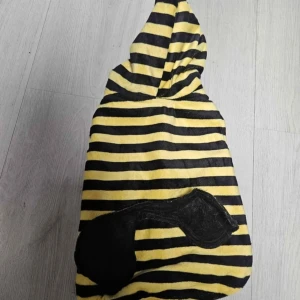 Randig jacka med huva i bumblebee-stil - Gul och svart randig jacka med huva för hunden och dekorativt svart märke på ryggen. Jackan är tillverkad i mjuk polyester och har öppningar för framben. Perfekt för dig som vill sticka ut med en färgglad och lekfull look.