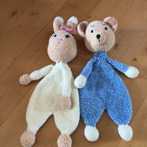 Handgjorda virkade gosedjur kanin och björn - Två mjuka virkade gosedjur, en kanin med vit kropp, beige detaljer och rosa rosett samt en björn med blå kropp och beige detaljer. Perfekta som kramdjur eller dekoration. Båda har broderade ansikten och är tillverkade i mjukt garn.