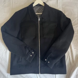 Svart jacka med dragkedja - Säljer en stilren svart jacka med silverfärgad dragkedja framtill och två bröstfickor med lock. Jackan har klassisk krage och knappar vid ärmsluten. Passar perfekt till dig som gillar enkel och clean stil. Bomullsblanding 