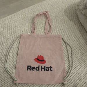 Ljusrosa tygpåse från Red Hat - Säljer en ljusrosa tygpåse från Red Hat med tryckt logga och text på framsidan. Väskan har både handtag och snören för att kunna bäras som ryggsäck. Tillverkad i återvunnen bomull.
