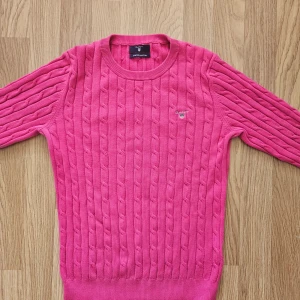 Rosa stickad tröja från Gant - Säljer en snygg rosa stickad tröja från Gant i stretchig bomull. Tröjan har ett klassiskt kabelstickat mönster och rund halsringning. Perfekt för en stilren look! Har använd få gånger men är i bra skick.
