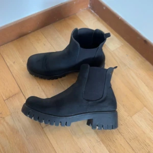 Svarta chelsea boots med grov sula - Säljer ett par svarta chelsea boots med rejäl, grovmönstrad sula. Skorna har elastiska paneler på sidorna och är tillverkade i syntetmaterial. Perfekta för en edgy och stilren look.