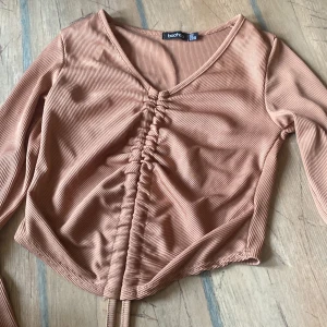 Beigefärgad ribbad croppad topp från Boohoo - Trendig beigefärgad croppad topp från Boohoo med ribbad struktur och v-ringning. Toppen har långa ärmar och snygg knytdetalj framtill som ger en rynkad effekt. Perfekt för att skapa en cool och avslappnad look.