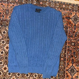 Blå stickad tröja från East West - Säljer en snygg blå stickad tröja från East West. Tröjan har ett klassiskt kabelstickat mönster och långa ärmar. 