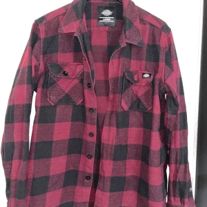 Röd och svart rutig skjorta från Dickies - Säljer en klassisk röd och svart rutig skjorta från Dickies. Skjortan har långa ärmar och knappar framtill samt på bröstfickorna. Perfekt för en avslappnad stil med en touch av retro. Tillverkad i bomull för en bekväm känsla.storlek Large