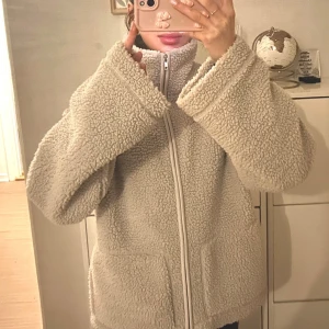 Beige teddyjacka med dragkedja - Mysig beige teddyjacka med dragkedja framtill. Perfekt för kyliga dagar med sin fluffiga och varma design. Jackan har långa ärmar och en hög krage för extra värme.