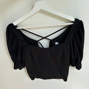 Svart topp med puffärmar och snörning - Säljer en svart croptop i satin med puffärmar och snygga snörningsdetaljer i ryggen. Toppen har elastiska ärmslut och tunna band som korsas i ryggen för en trendig look. 
