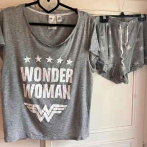 Grå Wonder Woman pyjamas med t-shirt och shorts från H&M - Säljer ett grått set med Wonder Woman-tema från H&M. T-shirten har ett tryck med texten 'Wonder Woman' och stjärnor, medan shortsen har ett stjärnmönster. Setet är tillverkat av bomull och viskos, vilket gör det mjukt och bekvämt. Tecken på användning finns, se bilder.
