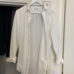 Vit skjorta - Säljer en klassisk vit skjorta från Lacoste. Skjortan har långa ärmar och knappar framtill. Perfekt för en stilren look. Storlek S, perfekt till sommaren och alla tillfällen!!