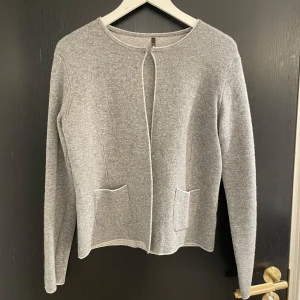 Grå kofta från Martty - Säljer en stilren grå kofta från Martty. Den har en enkel design med rund hals och två fickor framtill. Perfekt för lager-på-lager under kyligare dagar. 